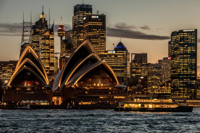 © Bloomberg. The Sydney Opera House, foreground, and buildings in the financial district stand illuminated at dusk in Sydney, Australia, on Friday, Sept. 29, 2017. A bungled transition from coal to clean energy has left resource-rich Australia with an unwanted crown: the highest power prices in the world. 