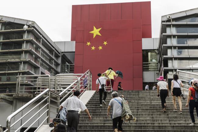 © Bloomberg. People walk up a flight of stairs as a Chinese flag is displayed at a border crossing facility in the Sha Tou Jiao Port of Shenzhen, China, on Friday, May 18, 2018. China’s growth this year shows no signs of slowing, despite rising tensions with the U.S. over trade and disappointing economic data in May, according to a Bloomberg survey. Photographer: Qilai Shen/Bloomberg