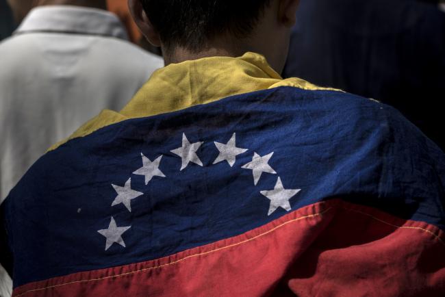 © Bloomberg. An attendee wears a Venezuelan flag during a rally with Juan Guaido, president of the National Assembly who swore himself in as the leader of Venezuela, not pictured, at Bolivar Square in Caracas, Venezuela. Photographer: Marcelo Perez del Carpio/Bloomberg