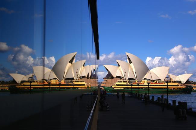 © Bloomberg. A ferry is reflected in a window as it passes the Sydney Opera House in Sydney, Australia, on Wednesday, May 15, 2019. Australia's economy has been weighed down by a retrenchment in household spending as property prices slump and slash personal wealth. An election Saturday is likely to see the opposition Labor party win power and lift spending further. 