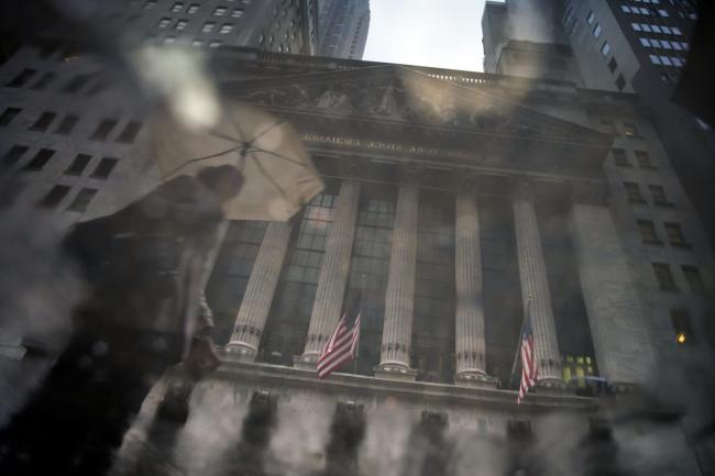 © Bloomberg. A pedestrian carrying an umbrella is reflected in a puddle in front of the New York Stock Exchange (NYSE) in New York, U.S., on Thursday, March 29, 2018. U.S. stocks climbed as markets moved toward the end of a tumultuous quarter on a high note. Equities in Europe also rose following a mixed session in Asia.