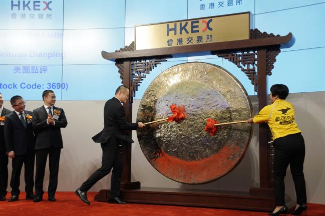 © Bloomberg. Wang Xing, chairman, chief executive officer and co-founder of Meituan Dianping, center, and Qiu Xuexue, one of the company's delivery workers, strike a gong during the company's listing ceremony at the Hong Kong Stock Exchange in Hong Kong, China, on Thursday, Sept. 20, 2018.  Photographer: Anthony Kwan/Bloomberg