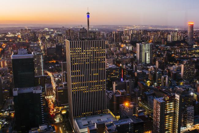 © Bloomberg. Commercial buildings and office property stand on the city skyline as night falls, as seen from the 50th floor of the Carlton Centre, in Johannesburg, South Africa, on Tuesday, July 17, 2018. South Africa's cash-strapped power utility, Eskom Holdings SOC Ltd., is 