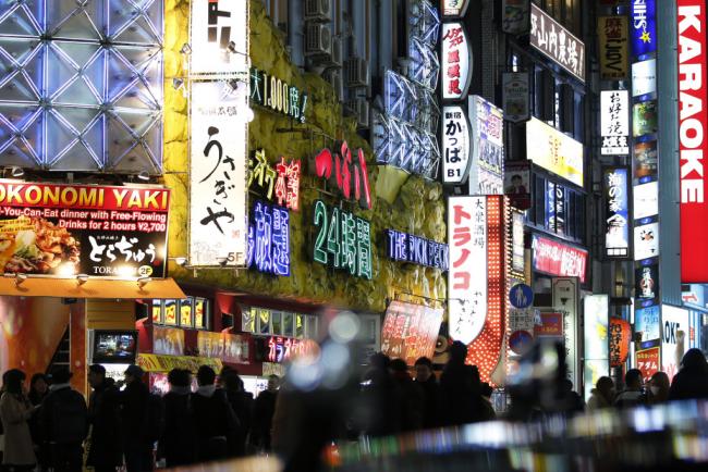 © Bloomberg. Pedestrians walk along a street illuminated with neon signs in Tokyo, Japan, on Thursday, Dec. 11, 2014. 