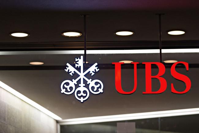 © Bloomberg. An illuminated sign hangs above an entrance to the UBS Group AG headquarters in Zurich, Switzerland, on Saturday, Dec. 24, 2016.  