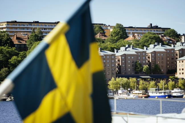 © Bloomberg. Residential housing blocks stand near the waterside beyond a Swedish national flag in the Soeder Maelarstrand area of Soedermalm in Stockholm, Sweden.