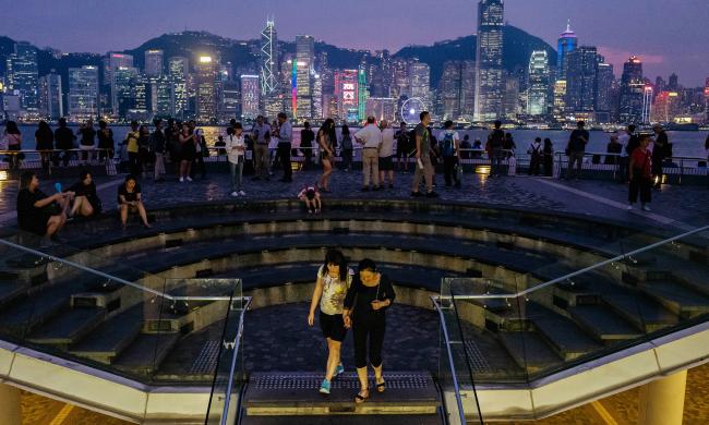 © Bloomberg. People look out from the waterfront in the Tsim Sha Tsui district as buildings across the Victoria Harbour stand illuminated at night in Hong Kong. Photographer: Anthony Kwan