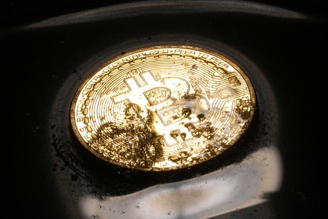 © Bloomberg. A coin representing Bitcoin cryptocurrency sits in a pool of melted ice crystals and water in this arranged photograph in London, U.K., on Wednesday, Feb. 7, 2018. Cryptocurrencies tracked by Coinmarketcap.com have lost more than $500 billion of market value since early January as governments clamped down, credit-card issuers halted purchases and investors grew increasingly concerned that last year’s meteoric rise in digital assets was unjustified. 