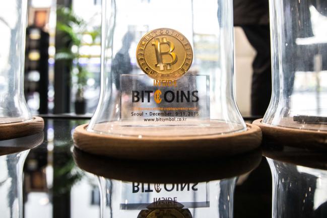 © Bloomberg. A gold colored bitcoin token sits on display under a glass cloche inside the offices of La Maison du Bitcoin bank in Paris, France.