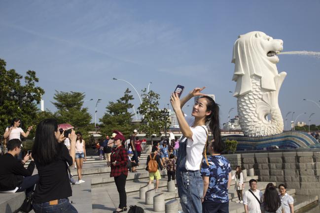 © Bloomberg. A tourist poses for a selfie photograph in front of the Merlion statue at the Marina Bay waterfront in Singapore, on Friday, Feb. 16, 2018. Singapore’s dollar is little changed near a three-week high amid thin trading with many Asian markets closed for Lunar New Year holidays. The central bank will announce the amount of 30-year bonds to be auctioned next week in the only planned sale of this tenor in 2018. Photographer: Ore Huiying/Bloomberg