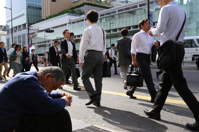 © Bloomberg. Morning commuters walk past a man sitting outside Shinjuku Station in Tokyo, Japan, on Tuesday, May 15, 2018. Japan is scheduled to release its first-quarter gross domestic product (GDP) figures on May 16. Photographer: Takaaki Iwabu/Bloomberg