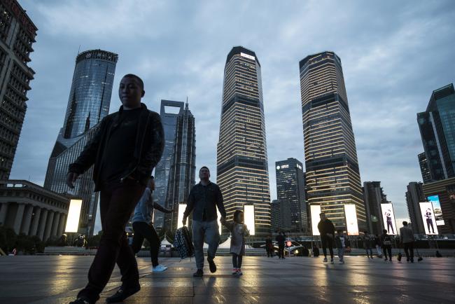 © Bloomberg. Pedestrians walk past commercial buildings illuminated at dusk in the Lujiazui Financial District in Shanghai, China.