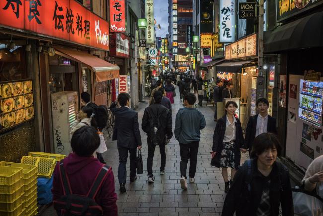 © Bloomberg. People walk past stores in the Shinjuku District of Tokyo, Japan, on Saturday, April 14, 2018. Japan's headline inflation measure is expected to rise to 1.1 percent this year and in 2019, from 0.5 percent in 2017, the International Monetary Fund said in its World Economic Outlook on April 17. 