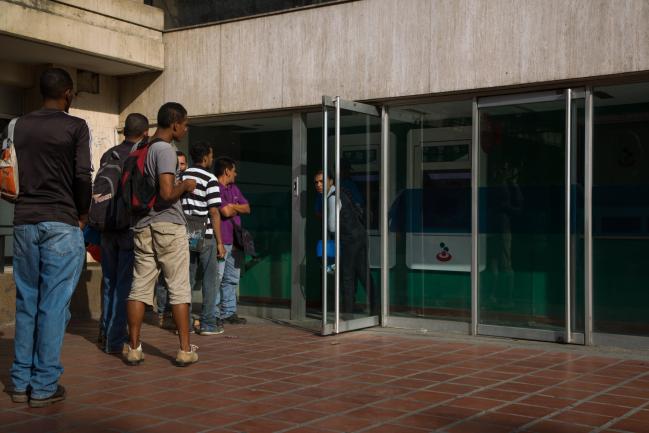 © Bloomberg. Customers stand in line to use automatic teller machines (ATM) at a Banesco Banco Universal CA bank branch in Caracas, Venezuela, on Thursday, May 3, 2018. Venezuela's chief prosecutor ordered the arrest of 11 high-ranking executives from the nation’s largest privately-held bank, Banesco Universal, as the government struggles to bring hyperinflation under control. Photographer: Wil Riera/Bloomberg