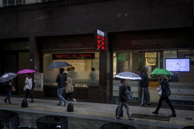 © Bloomberg. Pedestrians carry umbrellas while walking past a currency exchange house in Buenos Aires, Argentina, on Thursday, May 10, 2018. Argentine investors are shifting their focus to Tuesday's central bank auction of notes as a key test for President Mauricio Macri after the peso had its worst week since he came to power in 2015. 