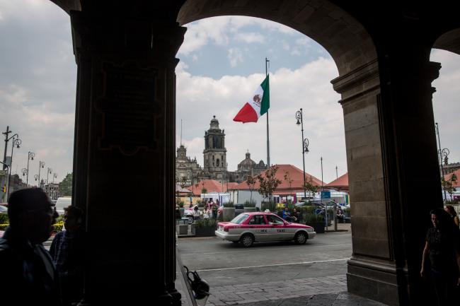 © Bloomberg. A taxi cab passes in front of the Mexican flag flying at the Plaza de la Constitucion (Zocalo) in Mexico City, Mexico, on Friday, April 13, 2018. 