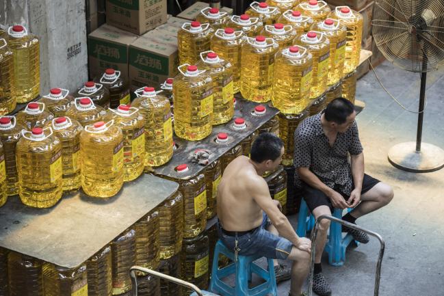 © Bloomberg. Two man sit talking next to stacked containers of soybean oil at a wholesale market in Shanghai, China, on Friday, July 13, 2018. China is scheduled to release gross domestic product (GDP) figures on July 16. 