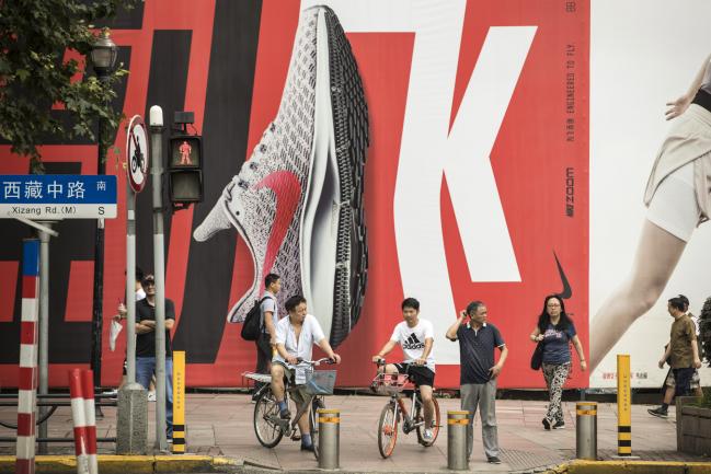© Bloomberg. Pedestrians and cyclists wait at a traffic signal in front of an advertising hoarding for Nike Inc. in Shanghai, China, on Friday, July 6, 2018. As a trade war looms, one of Chinese President Xi Jinping's biggest weapons could be boycotts of American brands by his country's legion of consumers. Photographer: Qilai Shen/Bloomberg