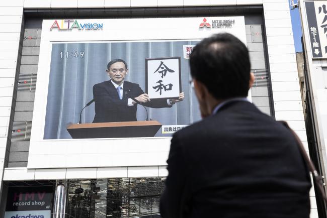 © Bloomberg. A pedestrian watches a news broadcast of Japan's Chief Cabinet Secretary Yoshihide Suga unveiling the name of Japan's next imperial era 