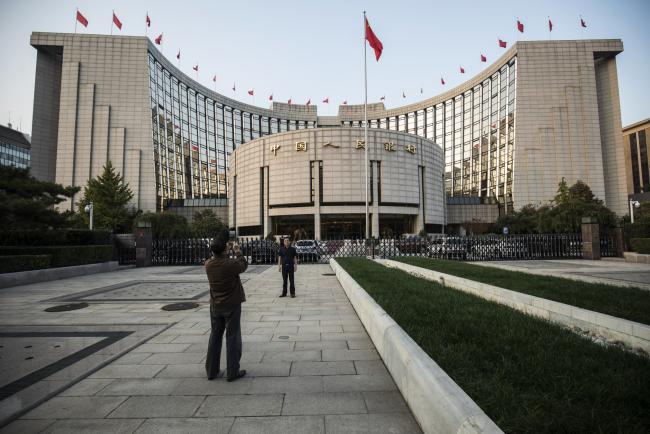 © Bloomberg. A man poses for a photograph in front of the People's Bank of China headquarters in Beijing. 
