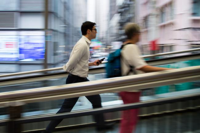 © Bloomberg. A man holding a smartphone walks along a moving walkway in the Central district of Hong Kong, China, on Thursday, Sept. 29, 2016. Hong Kong faces growing dissatisfaction over China's stewardship of the former British colony, with pro-democracy protests blocking city streets for weeks in 2014 and the subsequent emergence of new groups advocating independence.