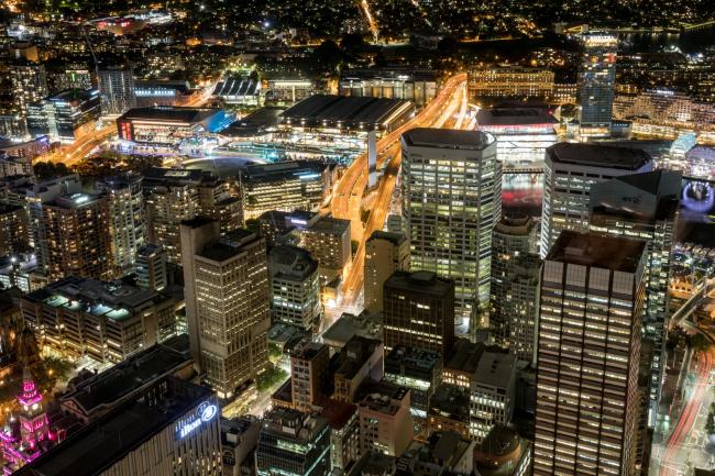 © Bloomberg. Buildings in the financial district stand illuminated at night in Sydney, Australia, on Friday, Sept. 29, 2017. A bungled transition from coal to clean energy has left resource-rich Australia with an unwanted crown: the highest power prices in the world.