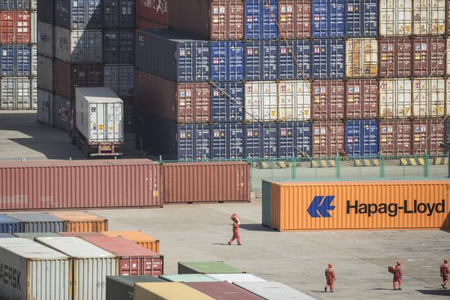 © Bloomberg. Workers walk past containers stacked at the Yangshan Deep Water Port in Shanghai, China, on Tuesday, July 10, 2018. China told companies to boost imports of goods from soybeans to seafood and automobiles from countries other than the U.S. after trade tensions between the world's two biggest economies escalated into a tariff war last week. 