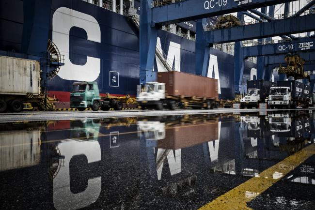 © Bloomberg. Trucks are reflected in a puddle as they wait in line to unload their containers onto CMA CGM SA's Benjamin Franklin container ship docked at the Guangzhou Nansha Container Port in Guangzhou, China, on Monday, Feb. 1, 2016. 