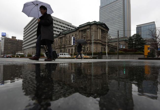 © Bloomberg. Pedestrians holding umbrellas while walking past the Bank of Japan (BOJ) headquarters are reflected in a puddle in Tokyo, Japan, on Friday, Jan. 29, 2016. Haruhiko Kuroda, BOJ governor, sprung another surprise on investors Friday, adopting a negative interest-rate strategy to spur banks to lend in the face of a weakening economy.