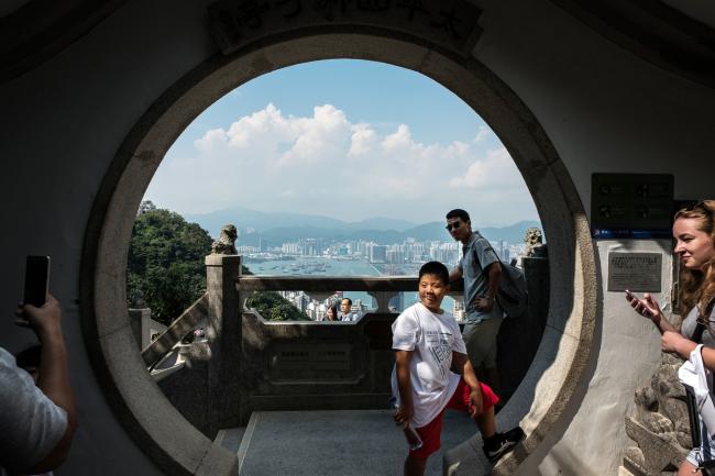 © Bloomberg. Tourists pose for a photograph at Victoria Peak in Hong Kong, China, on Monday, Sept. 11, 2017. Hong Kong stocks fluctuated on Sept. 12 as automakers extended gains driven by China's plan to phase out fossil-fuel vehicles, while banks and property companies weighed on the benchmark index.