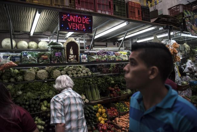 © Bloomberg. Customers browse a vegetable stand in the Chacao Municipal market in Caracas, Venezuela, on Saturday, April 14, 2018. As widespread food shortages continue, market vendors are selling small bagged portions of groceries; through February, the cost of a basket of goods needed monthly to support an average family of 5 in Venezuela rose 3,464% from a year prior. Photographer: Carlos Becerra/Bloomberg