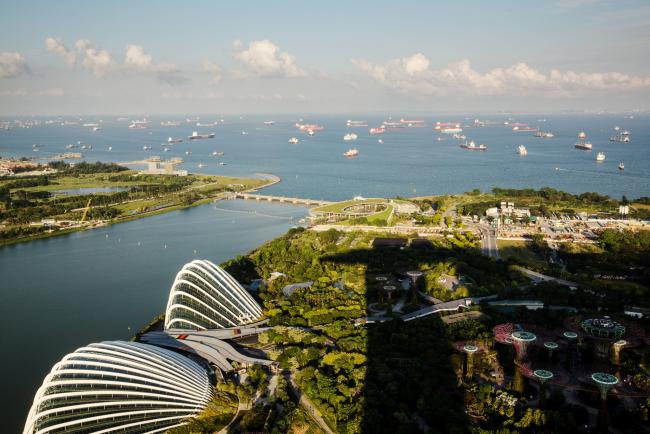 © Bloomberg. The Marina Bay Sands hotel and casino casts a shadow over the Gardens by the Bay botanical park as container ships are seen in the Singapore Strait in Singapore, on Sunday, June 4, 2017. After two years of below-par growth, economists and even Singapore's government are becoming more positive on the city-state's outlook. While it's not boom time yet, the consensus is that 2017 growth will come in higher than last year’s 2 percent. Photographer: Sanjit Das/Bloomberg