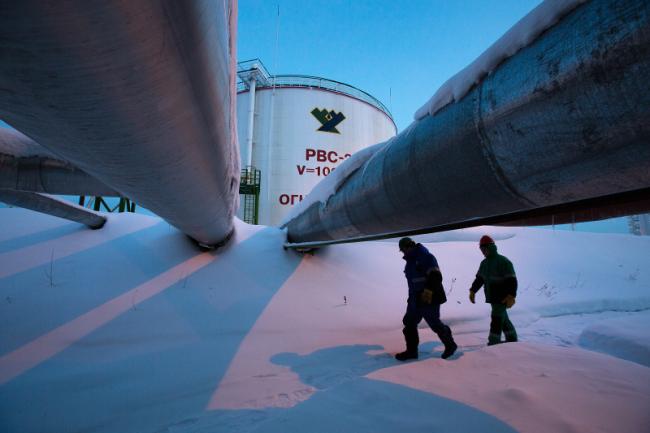 © Bloomberg. Employees pass beneath pipes leading to oil storage tanks at the central processing plant for oil and gas at the Salym Petroleum Development oil fields near the Bazhenov shale formation in Salym, Russia.