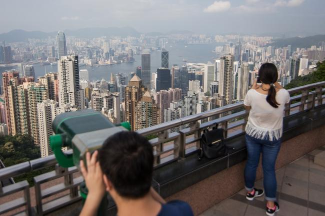 © Bloomberg. A tourist, left, uses binoculars to look at the view from Victoria Peak as commercial and residential buildings stand in the distance in Hong Kong, China, on Tuesday, April 28, 2015. Total visitors to Hong Kong, including those from China, fell 12.4 percent during the April 3 to 7 Ching Ming and Easter holidays, the government said, with Chief Executive Leung Chun-ying describing the decline as a 