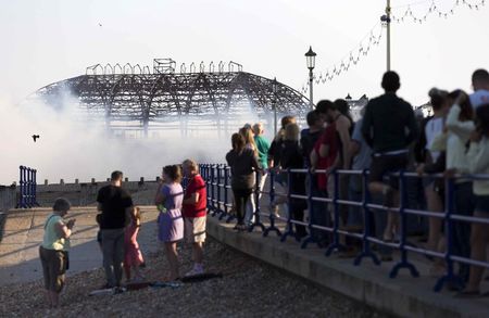 © Reuters. Members of the public watch as fire fighters from the East Sussex Fire and Rescue Service work to dampen down flames following a fire on the pier in Eastbourne