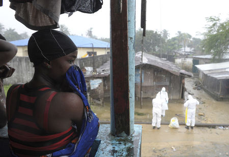 © Reuters. Liberian woman stands as health workers wearing protective clothing prepare to carry an abandoned dead body presenting with Ebola symptoms at Duwala market in Monrovia