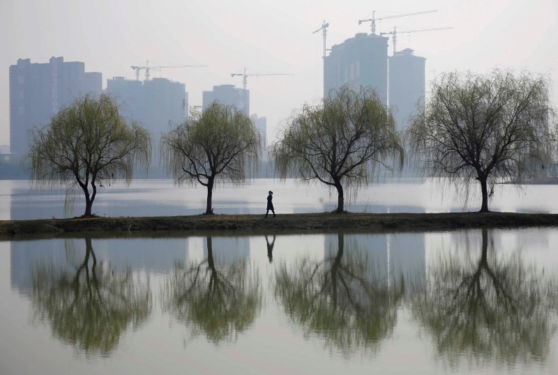 © Reuters. A woman walks past trees reflected on a lake in front of a construction site of a residential compound on a hazy day in Wuhan