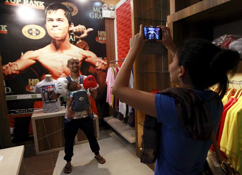 © Reuters. Woman uses a mobile phone to take pictures of her husband posing in front of a poster of boxer Manny Pacquiao of the Philippines on display at a mall in Manila 