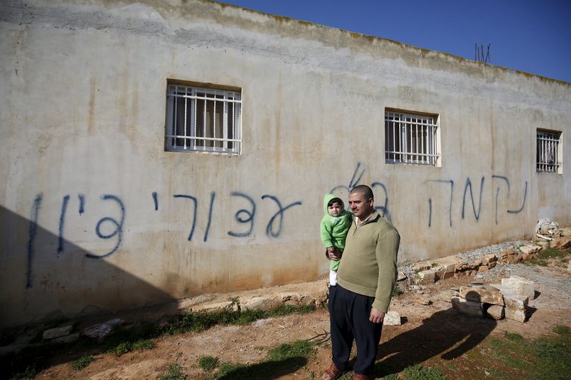 © Reuters. A Palestinian man holds his son as he stands in front of Hebrew graffiti sprayed on the wall of a Palestinian home in what Israeli police believe was an attack by Jewish militants in the village of Beitillu near the West Bank city of Ramallah