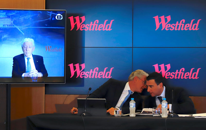 © Reuters. Westfield Chairman and co-founder Frank Lowy appears on a screen via video-link as his son Steven Lowy talks with Elliot Rusanow, Chief Financial Officer of Westfield, during a media conference in Sydney