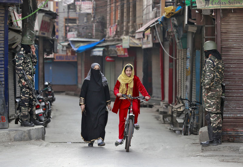 © Reuters. FILE PHOTO: A Kashmir girl rides her bike past Indian security force personnel standing guard in front closed shops in a street in Srinagar