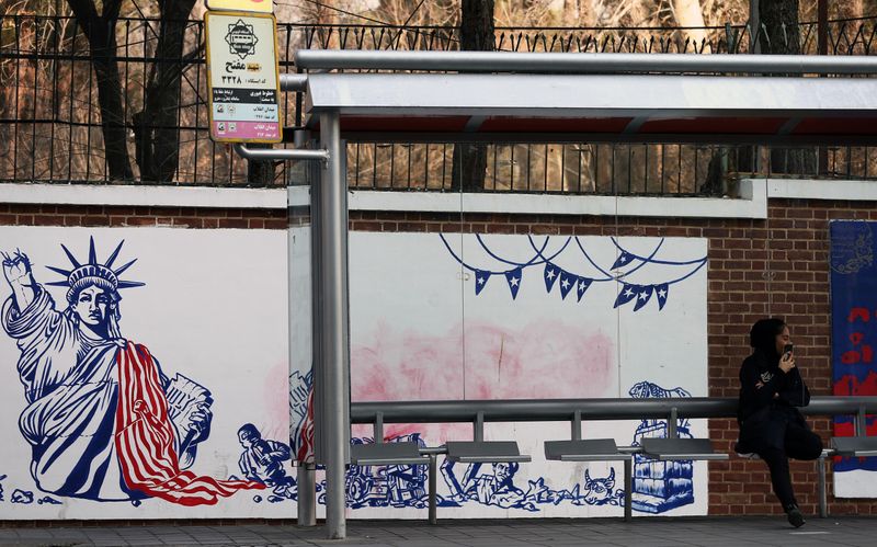© Reuters. A woman sits at a bus stop in front of an Anti US mural on the former US embassy's wall in Tehran