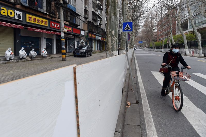 &copy; Reuters. Woman wearing a face mask rides a shared bicycle past workers in protective suits who are resting in front of closed shops sealed off from the road, in Wuhan