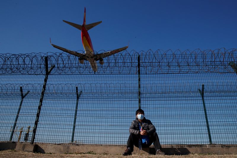 © Reuters. A man wearing a protective mask sits at a fence surrounding Beijing Capital International as a plane lands in Beijing as the country is hit by an outbreak of the novel coronavirus