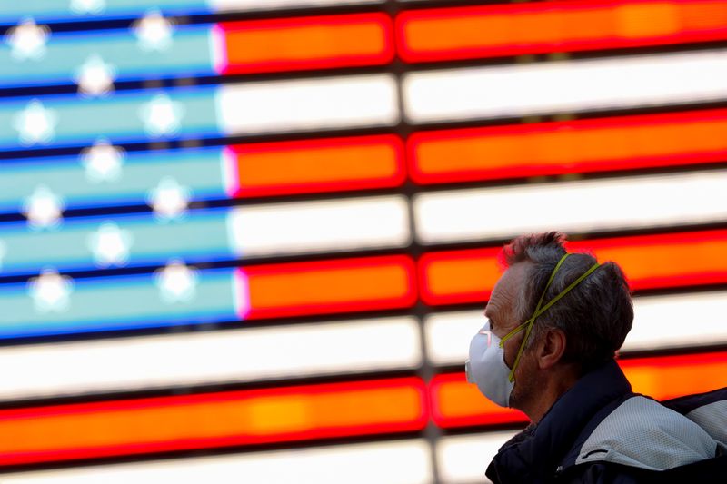 &copy; Reuters. Un uomo in maschera chirurgica cammina accanto a una bandiera degli Stati Uniti a Manhattan