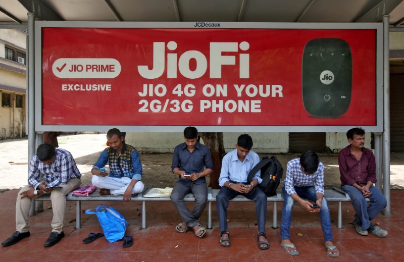 © Reuters. FILE PHOTO: Commuters use their mobile phones as they wait at a bus stop with an advertisement of Reliance Industries' Jio telecoms unit, in Mumbai