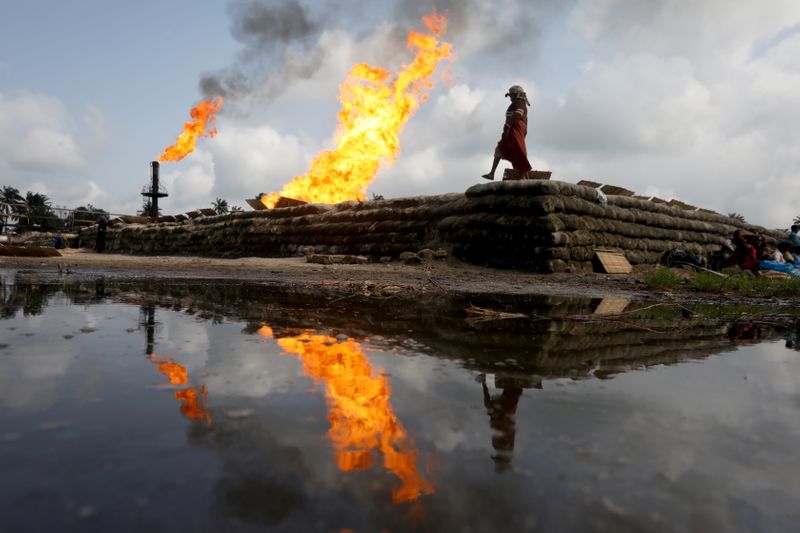 © Reuters. A reflection of two gas flaring furnaces and a woman walking on sand barriers is seen in the pool of oil-smeared water at a flow station in Ughelli, Delta State