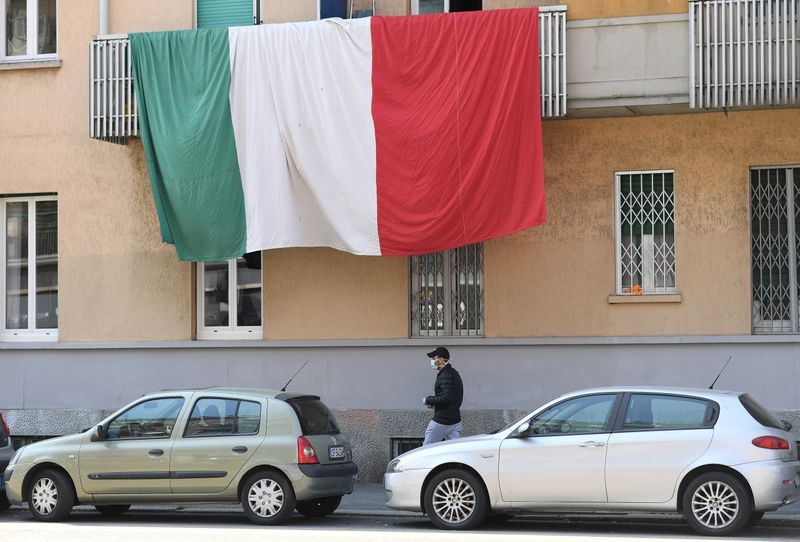 &copy; Reuters. Un uomo con una mascherina cammina sotto a una bandiera italiana su un balcone a Milano