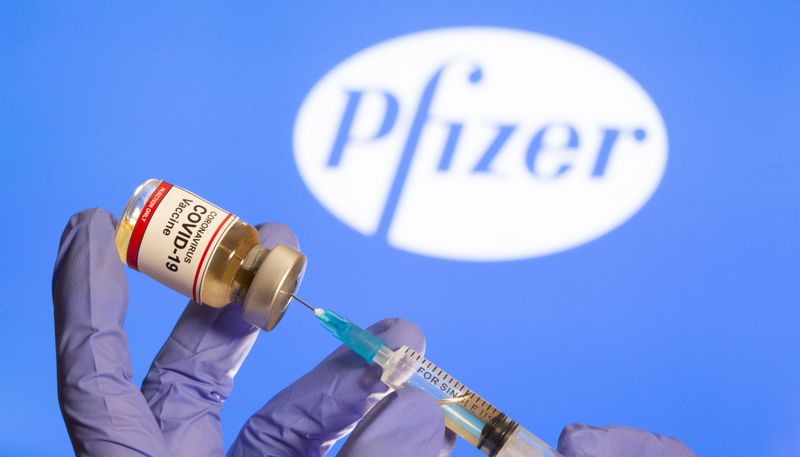 &copy; Reuters. A woman holds a small bottle labeled with a &quot;Coronavirus COVID-19 Vaccine&quot; sticker and a medical syringe in front of displayed Pfizer logo in this illustration