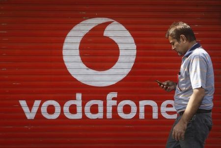 © Reuters. A man checks his mobile phone as he walks past a shop displaying the Vodafone logo on its shutter in Mumbai